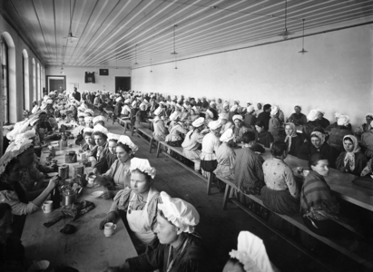 Women workers in a factory warehouse take a dinner break, supervised by a few male overseers (b/w photo)