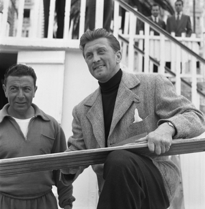 American actor Kirk Douglas, wearing a blazer and a neckturtle sweater, portrayed while fixing a pair of waterskis with sandpaper, a man smiling behind him, Venice 1953