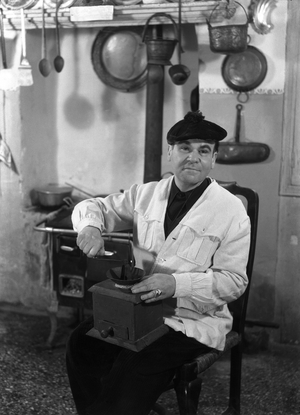Italian painter Filippo De Pisis, sitting on a chair in the kitchen, wearing a white shirt and a velvet beret and holding a coffee grinder, Venice, 1946