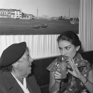 American gossip columnist Elsa Maxwell, wearing a beret and chatting with Maria Callas Meneghini, wearing a floral dress, pearl earrings and holding a drink, a painting with gondolas and St. Mark's Basin on the wall behind them during the Movie Festival, Biennale, Venice 1957