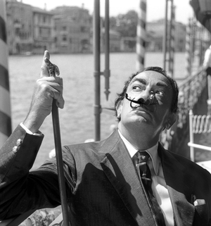 Spanish painter Salvador Dali', wearing a shantung blazer and a silk tie, holdind a walking-stick, with a small toy gondola as a moustache, sitting beside the Canal on a cafe terrace, Venice 1961