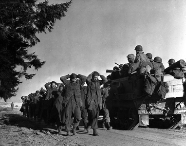 WWII:Near Bastogne (Belgium) 27 December 1944: As a half-track goes online, a 4th DB US military policeman brings German soldiers prisoners back