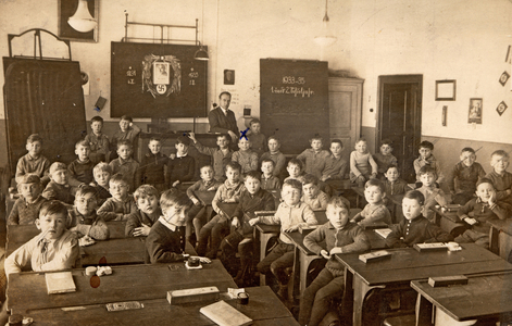 Photograph of a class in Germany under the Nazi regime in 1933. Cross ranges are hung on the blackboard and wall. The professor is standing at the back of the room.