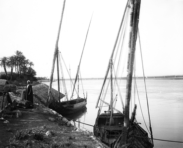 Egypt, Upper Egypt, Aswan: Cook cruise, village on the edge of the Nile, on the stone bank, near a large town and at the terminal of a railway, two felouques at the dock await their loads - At the bottom a gigantic bridge, the Aswan dam, 1900