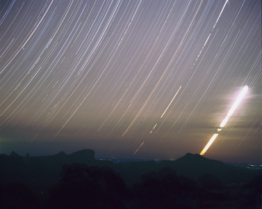 Queue d'étoiles - Moonset in the Warrumbungles - Photographic pose of several hours on the stars and the crescent moon. The picture here was made on one partially cloudy night at Siding Spring. In the foreground are the distinctive shapes of the Warrumbungle mountains in outback New South Wales. The exposure, on 400 ISO transparency film, was begun at the end of astronomical twilight, when the sun has descended 18 degrees below the horizon. There was a crescent moon and cloud around the south - western horizon. The exposure continued for several hours after the moon had set, and breaks in the star trails reveal that cloud had come and gone during the night
