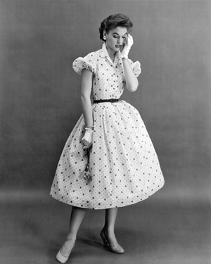 United States: c. 1956
A woman modeling a luscious strawberry print dress on fine dimity with tiny puffed sleeves to give that ""little girl"" look that is so fashionable today. Maker Nali-Bee also adds a touch of glitter with rhinestone buttons.