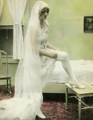 United States: c. 1925 - A hand tinted photograph of a nervous looking bride taking off her stockings and titled, ""The Bride Retires""