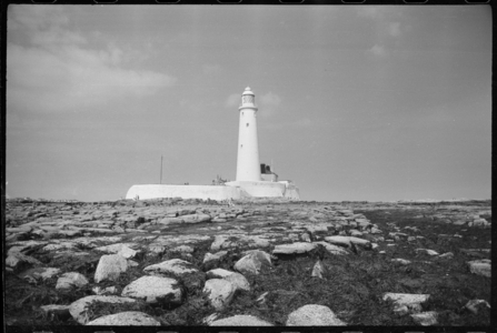 An exterior view of the 19th century lighthouse, showing the 126ft tall tower with a partial view of the Keeper's Cottage to the right, and a stone boundary wall surrounding the complex, seen from the east, St Marys Lighthouse, Whitley Bay, North Tyneside, North Tyneside, UK, c. 1955-c.1980 (b/w photo)