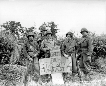 Five U.S. soldiers standing in front of a hedge, holding a cross-shaped sign with a text in German saying: "Soldaten ergebt euch, ihr seid umringt" ("Soldiers surrender, you are surrounded"), northwest of Saint- Lô , direction of Pont-Hébert and Hauts-Vents, Normandy, France, 14th July 1944 (b/w photo)