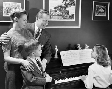 Rear view of girl playing piano with family with another kid watching