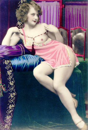A woman sitting in front of two small mirrors, wearing a pink undershirt pulled down on one side to expose her breast, 1925 (tinted photo)