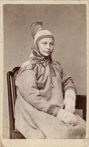 A half–length portrait of a seated woman wearing traditional Saami dress. She is dressed in a tunic, mittens, is wearing a hat with a high crest, and a triangular scarf tied around her neck. She also has a pendant hanging possibly from the cord that ties her hat under her chin, c.1870s (albumen print)
