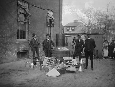 Onlookers watch as suited men stand in front of a large copper kettle still for making illegal liquor with boxes of bottles and funnels spread before them all for the manufacture of booze. (photo)
