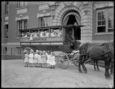 Small girls pose in and in front of a horse-drawn wagon, in front of an unidentified building, probably the Roman Catholic Orphan Asylum, Kingsbridge, Bronx, c.1910 (b/w photo)
