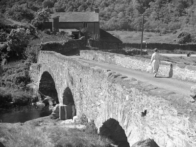 A woman walking across a bridge with multiple arches towards what appears to be a farm, 1930s-60s (b/w photo)