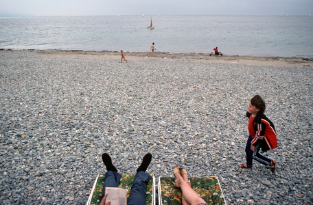 Couple resting and reading on stony beach at Criccieth seaside town, Llyn peninsula, Gwynedd, North Wales, 1983 (photo)