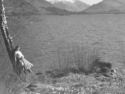 A view of a woman leaning against a tree alongside a lake, snow capped fells in the background, 1930s-60s (b/w photo)