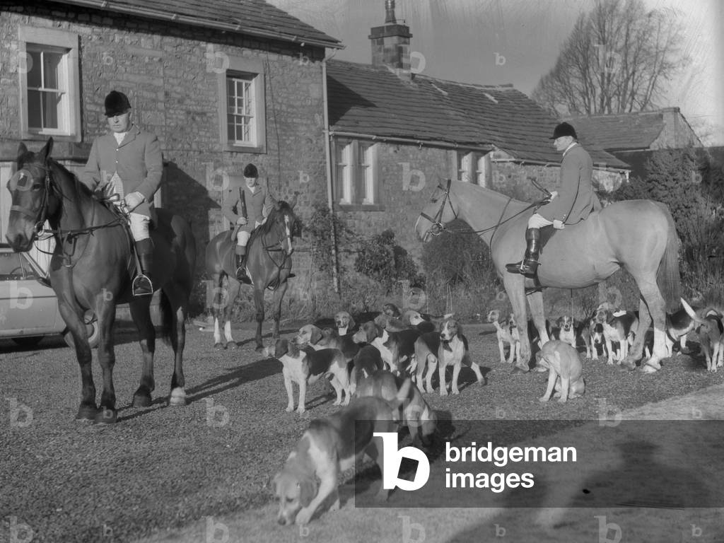 A view of huntsmen on horses and hunting hounds, all standing in front of a building, 1930s-60s (b/w photo)