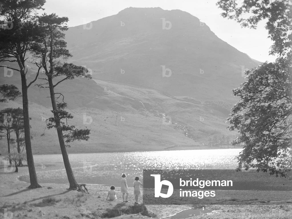 Three children standing on the shore of Buttermere with backs to camera looking at sunlit lake, 1930s-60s (b/w photo)