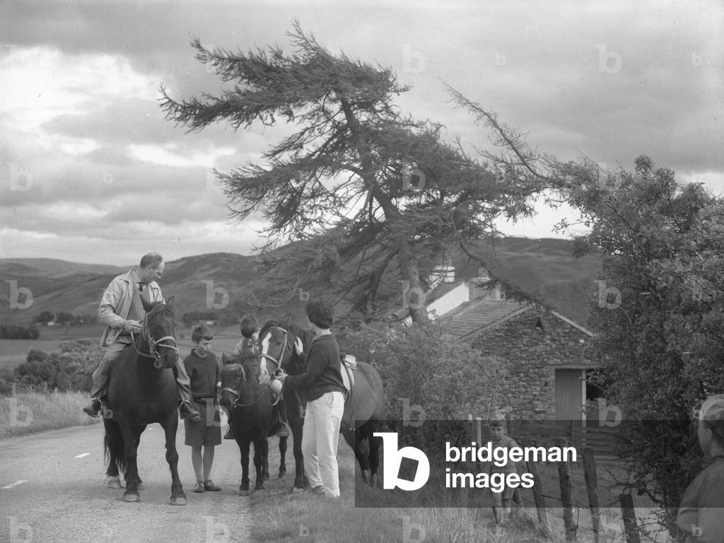 A view of a family stopped at the side of a road pony trekking, fells and building in the background, 1930s-60s (b/w photo)