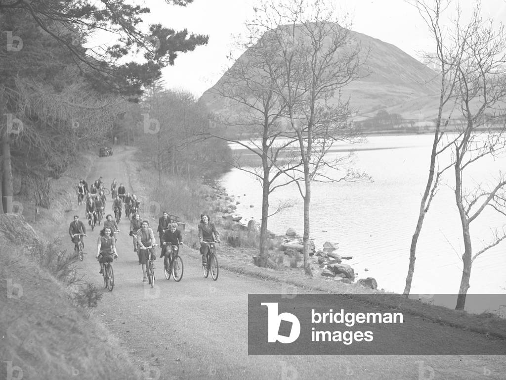 A view of a group of cyclists pedalling along a road at the side of a lake, fells in the background, 1930s-60s (b/w photo)