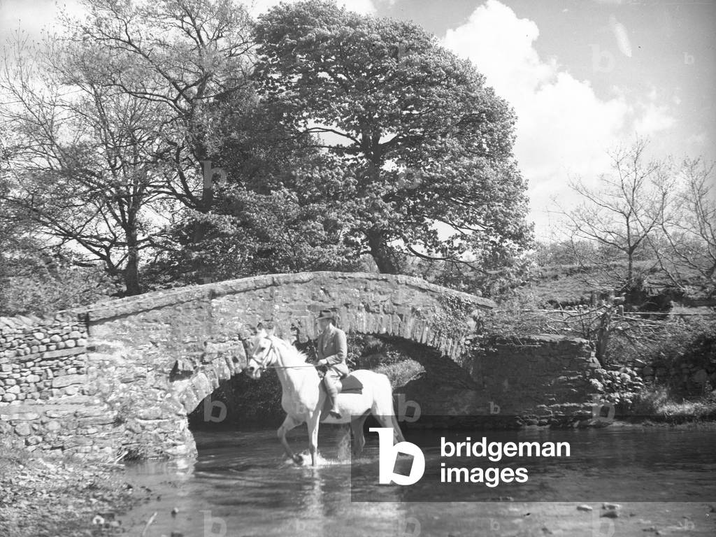 Johnty Wilson riding horse through shallow river next to bridge, 1930s-60s (b/w photo)