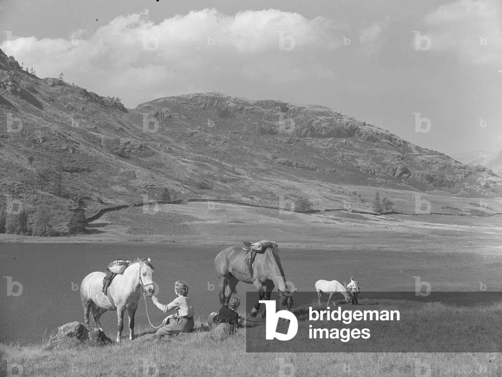 A view of women horse riders having a break with their horses near to a lake, fells in the background, 1930s-60s (b/w photo)