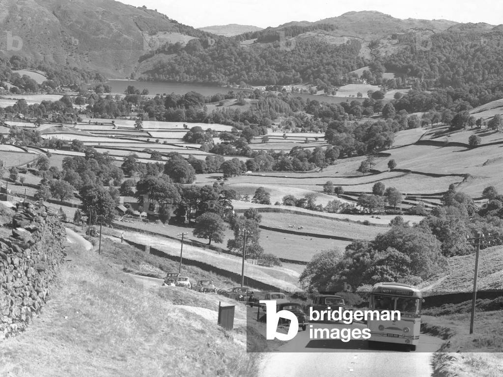 A view of a single decker bus followed by cars climbing up the old A591 toward Dunmail Pass (prior to improvements in 1970), farmland and lake Grasmere in the background, 1930s-60s (b/w photo)