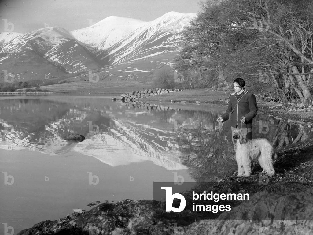 A man stands with his dog next to a lake, fells in background, 1930s-60s (b/w photo)