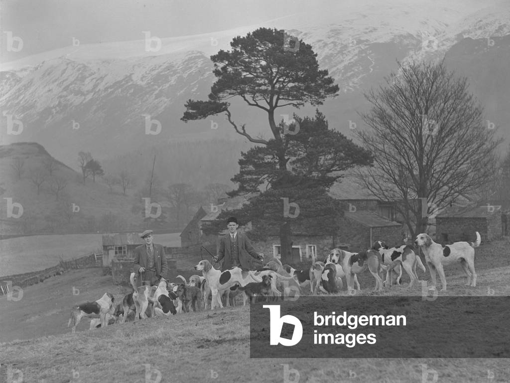 A group of hounds gather on a hillside with two men, fells covered in snow in background, 1930s-60s (b/w photo)