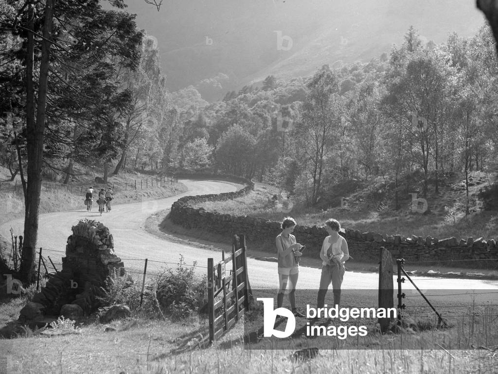 Two women stand talking in a gateway to a field, off a lane, 1930s-60s (b/w photo)