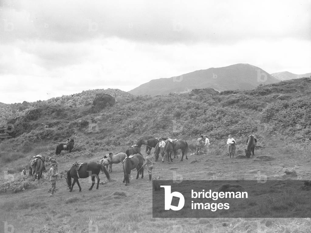 A group of horse riders and their horses taking a break on some grassland, fells in the background, 1930s-60s (b/w photo)