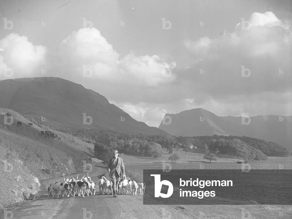 A man walks along a road with hounds at his feet, lake to the right and fells in background, 1930s-60s (b/w photo)