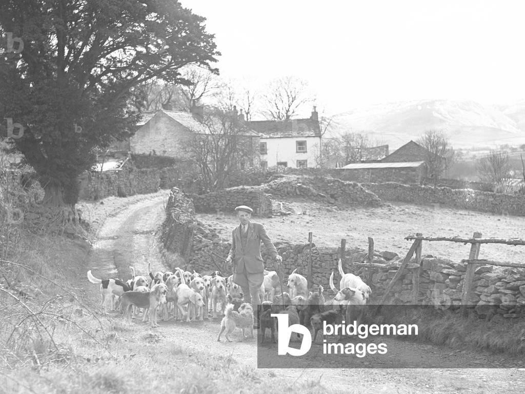 A man stands in a lane with hounds at his feet, building in the background (possibly on farm), 1930s-60s (b/w photo)