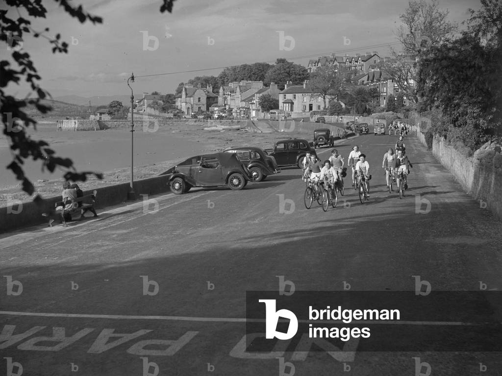 Group of boys cycling towards camera along front at Arnside with cars parked and Albion Hotel in background, 1930s-60s (b/w photo)