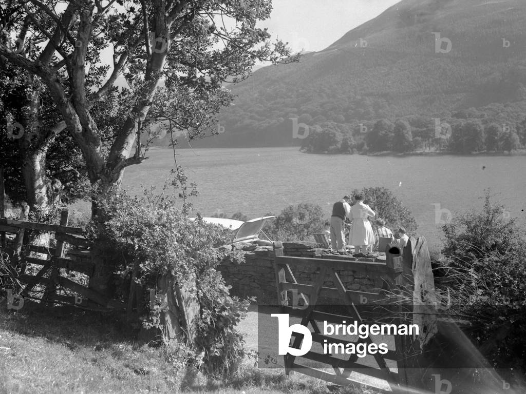 Family sat on a dry stone wall having a picnic overlooking lake, car parked up with boot open, 1930s-60s (b/w photo)