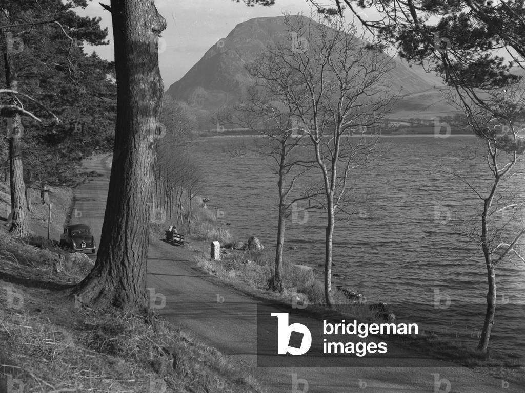 Two women sat on a bench overlooking a lake, car parked up on the lane, fells in background, 1930s-60s (b/w photo)