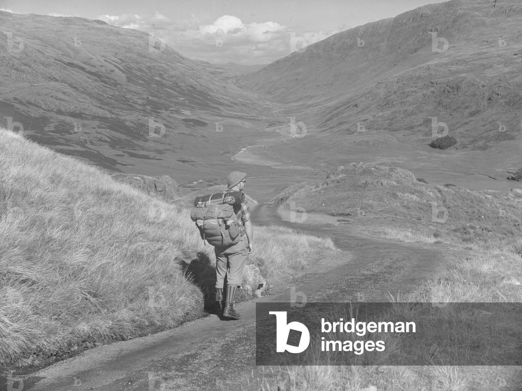 A man with a rucksack on his back walking down a road whilst looking out across a valley, 1930s-60s (b/w photo)