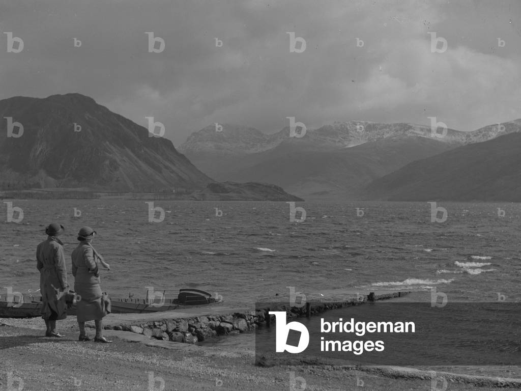 Two women stand next to a boat landing on a lake, fells in background, 1930s-60s (b/w photo)
