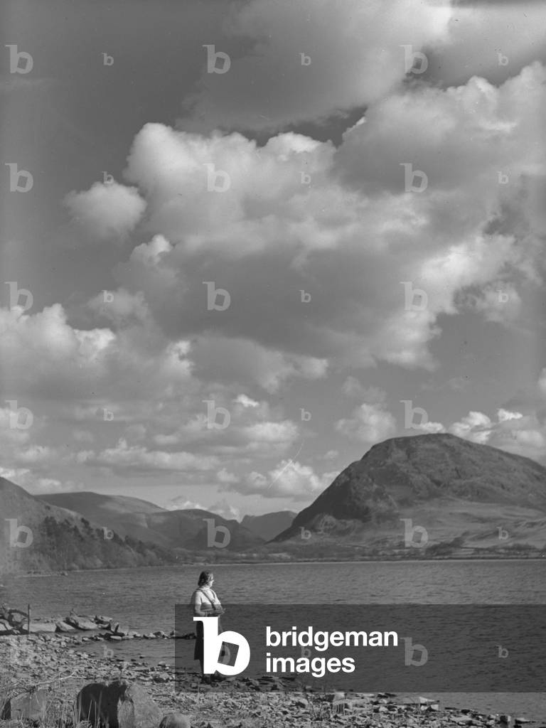 A view of a woman standing next to a lake with fells in the background, 1930s-60s (b/w photo)