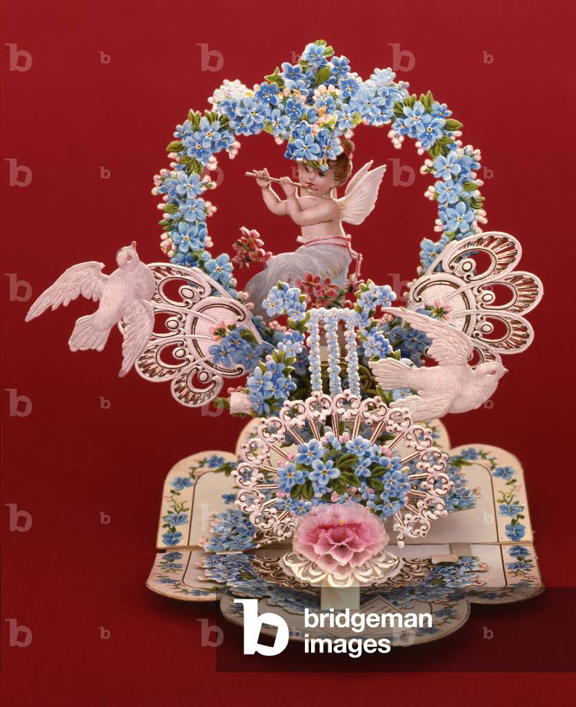 A pop-up card a winged cherub playing a flute under a heart shaped arch of forget-me-nots (chromolithograph on card)