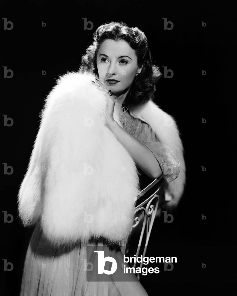 1940: American Screen Star Barbara Stanwyck (1907-1990) Wearing A Luxurious, White Fur Cape Over A Long Chiffon Evening Dress.