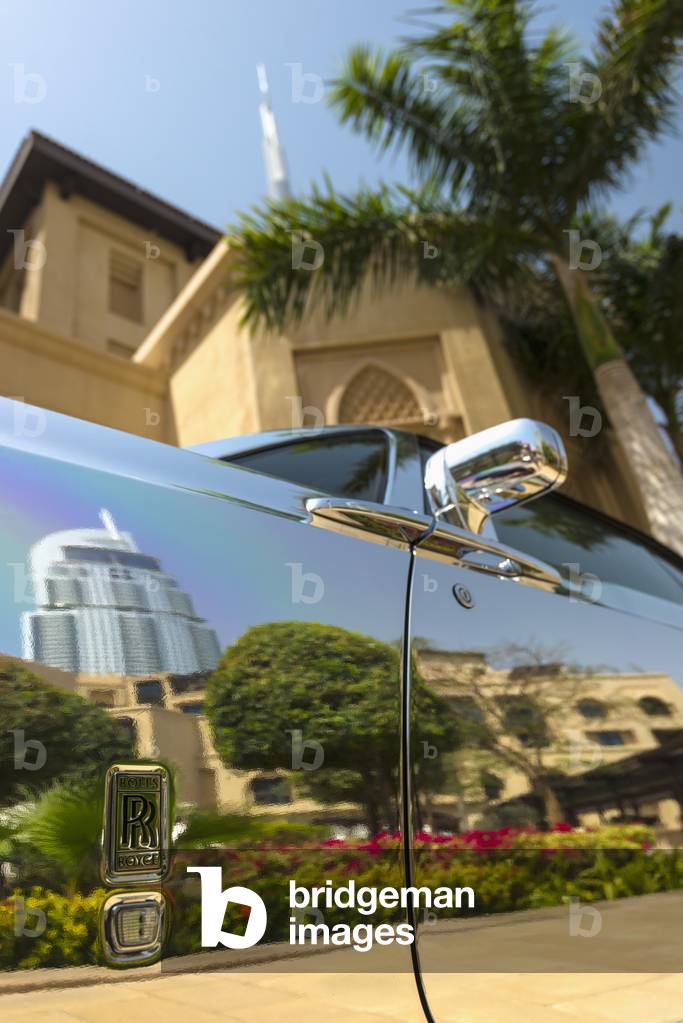 Detail of chrome plated Rolls Royce beneath palm tree and traditional arabic style hotel with the Burj Khalifa behind, Dubai, United Arab Emirates (photo)