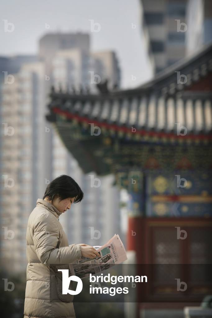 A Local Woman Reads A Newspaper, in The Background There Is A Contrast of Old  and New, A Traditional Chinese Structure Beside Modern Skyscrapers, Beijing, China (photo)