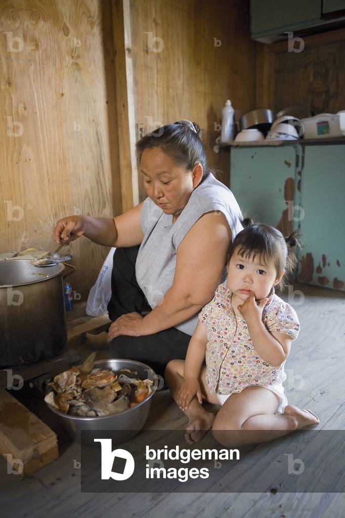 Native Yupik Woman W/Small Child Sits On Floor Of Cabin Cooking Salmon Akiachak Western Alaska (photo)