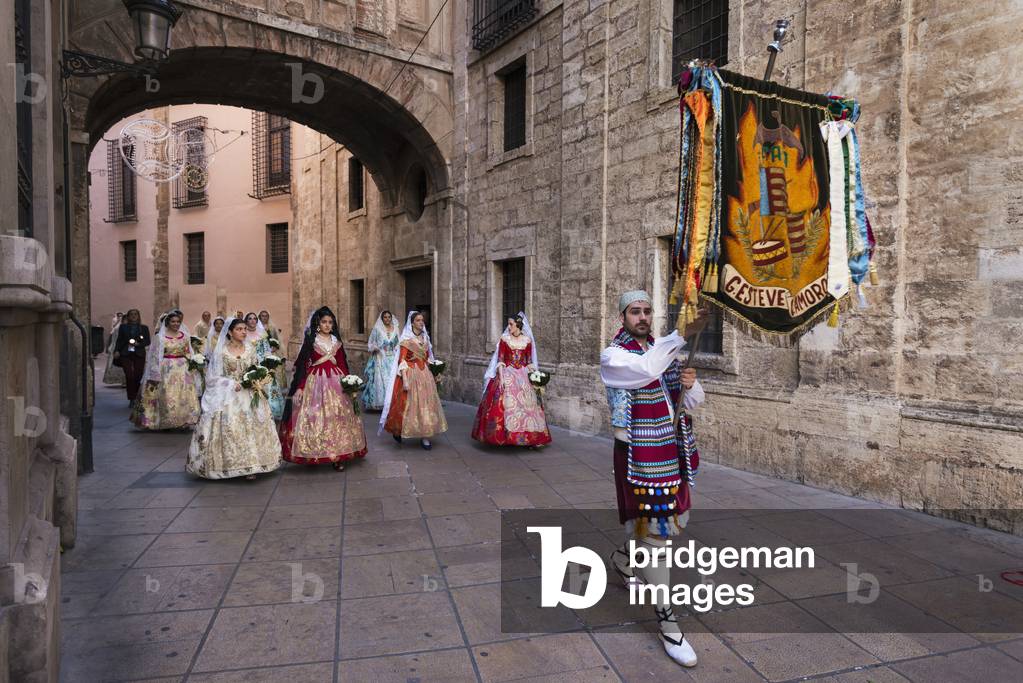 Man with Banner Leading Women Dressed up in Traditional Casal Faller Outfit with Flower Offering For Virgin De Los Desamparados at Fallas Festival, Valencia, Spain (photo)