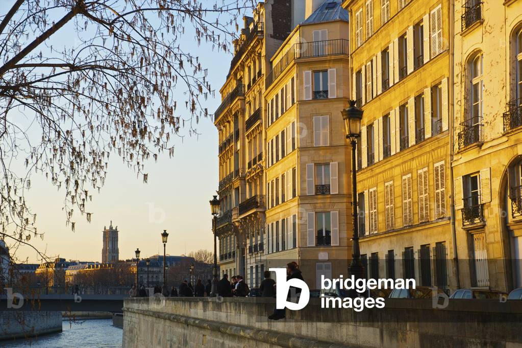 A couple sit together on a canal wall with sunset sunlight cast on the buildings, Paris, France (photo)