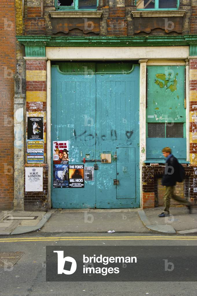 A pedestrian walks by a large blue door with posters, Shoreditch, London, England, UK  (photo)