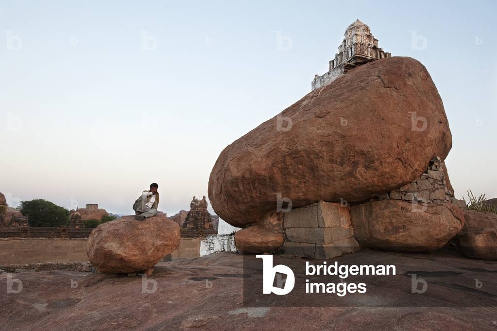 A structure sits atop a large boulder with a man sitting on a large rock next to it, Hampi, Karnataka, India (photo)