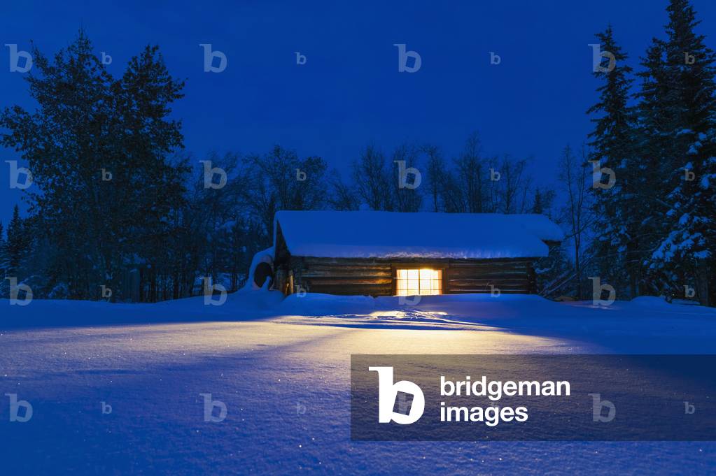 Warm light spills out of the window of a historic log cabin on the surrounding snow; Wiseman, Alaska, United States of America (photo)
